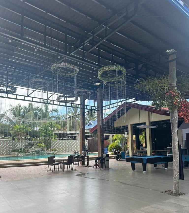High-roofed open-air pavilion with a billiard table, tables and hanging decorations, the pool beyond